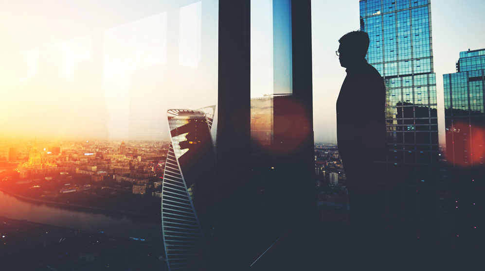 Bespectacled business man looking out over city at dusk.