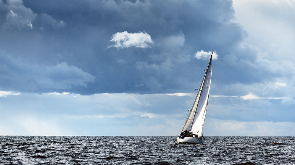 White sailboat sailing away against a dark sea and blue, clouded sky.