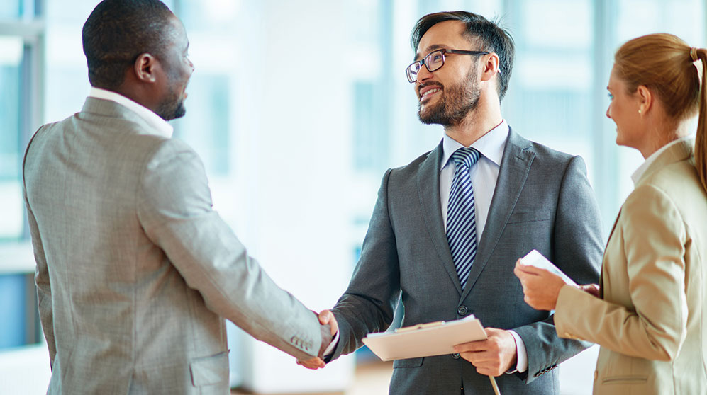 Two suited men shaking hands with a suited woman looking on smiling.